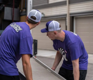 Two men in matching purple shirts and white caps bend over a long metal panel, working together outdoors near a loading dock.