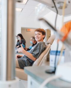 Boy smiling in a dental chair giving a thumbs up during a checkup; dental staff wearing masks in the background.