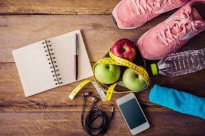 Flat lay of fitness planning items: notebook and pen, apples with a measuring tape, pink running shoes, water bottle, earphones, and a smartphone on a wooden floor.